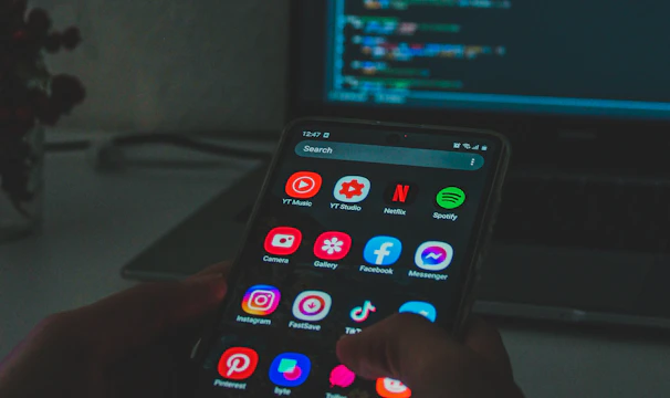 A person holds a smartphone displaying various colorful app icons, placed in front of a laptop screen showing a code editor with lines of code. The setting appears dimly lit, suggesting a focus on technology and coding.