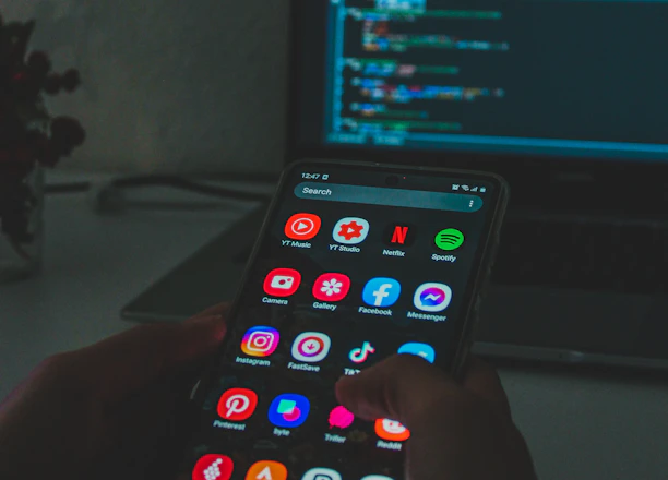 A person holds a smartphone displaying various colorful app icons, placed in front of a laptop screen showing a code editor with lines of code. The setting appears dimly lit, suggesting a focus on technology and coding.