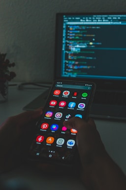 A person holds a smartphone displaying various colorful app icons, placed in front of a laptop screen showing a code editor with lines of code. The setting appears dimly lit, suggesting a focus on technology and coding.