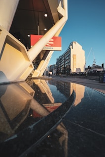 A modern architectural scene with reflective surfaces, featuring sleek, white structural elements and a glass facade capturing the reflection of nearby buildings. A construction crane is visible in the background, suggesting urban development. The atmosphere suggests a mixture of architectural precision and urban dynamism.