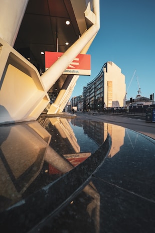 A modern architectural scene with reflective surfaces, featuring sleek, white structural elements and a glass facade capturing the reflection of nearby buildings. A construction crane is visible in the background, suggesting urban development. The atmosphere suggests a mixture of architectural precision and urban dynamism.