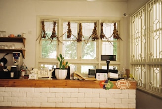 A cozy counter area featuring a rustic wooden counter with a white brick facade. The counter is adorned with various items including a small numbered stand, a plant in a white pot, and a digital display. Behind the counter, a window with hanging dried plants provides a view outside. Shelves to the left hold cups and utensils, adding to the homey atmosphere.
