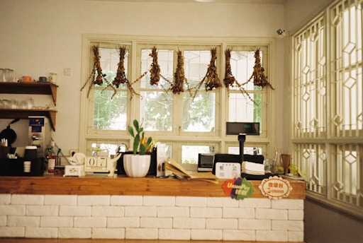 A cozy counter area featuring a rustic wooden counter with a white brick facade. The counter is adorned with various items including a small numbered stand, a plant in a white pot, and a digital display. Behind the counter, a window with hanging dried plants provides a view outside. Shelves to the left hold cups and utensils, adding to the homey atmosphere.