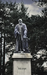 A statue of a historical figure standing on a pedestal surrounded by trees. The figure is dressed in traditional academic attire, including a long cloak, and holds a book under one arm. The word 'KELVIN' is inscribed on the pedestal.
