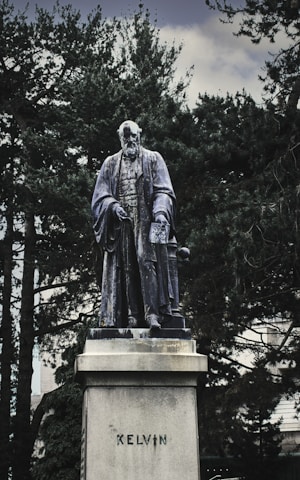 A statue of a historical figure standing on a pedestal surrounded by trees. The figure is dressed in traditional academic attire, including a long cloak, and holds a book under one arm. The word 'KELVIN' is inscribed on the pedestal.