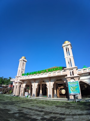 A grand mosque with two tall towers featuring gold domes and arches. The main entrance is adorned with a green sign displaying the mosque's name in large letters. The surrounding area includes a wide walkway with stone paths, surrounded by lush greenery. A bright blue sky stretches overhead, casting a clear and bright atmosphere.