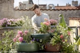 A person is tending to a rooftop garden filled with colorful flowers and plants grown in recycled tires. The background features urban architecture with brick walls and buildings, under a clear blue sky.