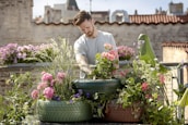 A person is tending to a rooftop garden filled with colorful flowers and plants grown in recycled tires. The background features urban architecture with brick walls and buildings, under a clear blue sky.