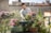 A person is tending to a rooftop garden filled with colorful flowers and plants grown in recycled tires. The background features urban architecture with brick walls and buildings, under a clear blue sky.