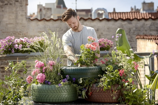 A person is tending to a rooftop garden filled with colorful flowers and plants grown in recycled tires. The background features urban architecture with brick walls and buildings, under a clear blue sky.