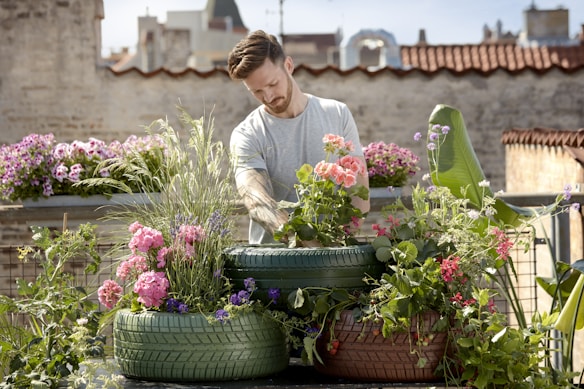 A person is tending to a rooftop garden filled with colorful flowers and plants grown in recycled tires. The background features urban architecture with brick walls and buildings, under a clear blue sky.