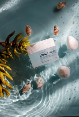 A jar labeled 'global oxygen' is floating in an aquatic setting surrounded by seashells, a white stone, and succulent plant leaves. The water creates gentle ripples around the objects, and sunlight casts soft shadows.