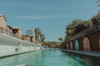 A long, narrow canal flanked by buildings with palm trees in the background. The left side features light brick buildings with white walls and railings, while the right side shows a darker structure adorned with colorful mural arches. The canal water is a vibrant green, reflecting the clear sky and surrounding architecture.