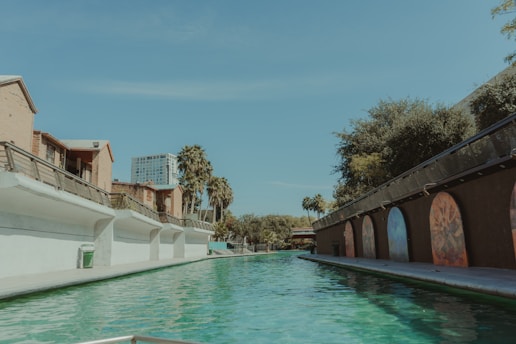 A long, narrow canal flanked by buildings with palm trees in the background. The left side features light brick buildings with white walls and railings, while the right side shows a darker structure adorned with colorful mural arches. The canal water is a vibrant green, reflecting the clear sky and surrounding architecture.