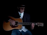 A person wearing a hat and sunglasses is playing an acoustic guitar. They are dressed in a dark blazer over a white shirt, with a turquoise bolo tie and blue jeans. The lighting is low, creating a moody and intimate atmosphere.
