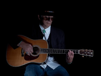 A person wearing a hat and sunglasses is playing an acoustic guitar. They are dressed in a dark blazer over a white shirt, with a turquoise bolo tie and blue jeans. The lighting is low, creating a moody and intimate atmosphere.