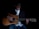 A person wearing a hat and sunglasses is playing an acoustic guitar. They are dressed in a dark blazer over a white shirt, with a turquoise bolo tie and blue jeans. The lighting is low, creating a moody and intimate atmosphere.
