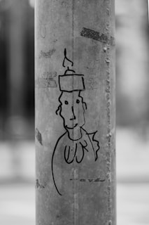 A black and white cartoon-style drawing on a concrete surface features a simplistic face with distinct eyes and a candle on top as part of the head. The drawing is outlined in black, and the surface has visible texture and some smudges.