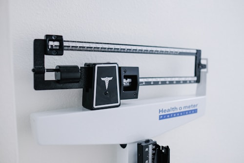 A professional mechanical doctor's scale with a white base and black measuring beam, featuring a caduceus symbol. The scale is positioned against a light-colored wall, indicating it is likely in a clinical or health-related environment.