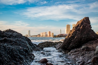 rocky shore with water waves near city buildings during daytime
