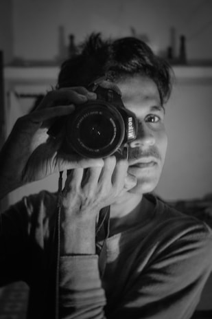 A person holding a DSLR camera close to their face, capturing their own image in a black and white setting. The focus is on the camera lens and their eye, creating a dramatic lighting effect with soft shadows.
