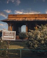 A traditional wooden structure with intricate carvings is seen against a partly cloudy sky. In the foreground, there is a sign indicating the direction to a cloakroom with text in both Hindi and English. Lush green bushes surround the area, and the sunlight casts warm tones over the scene.