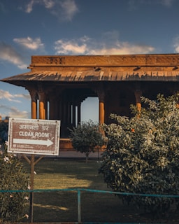 A traditional wooden structure with intricate carvings is seen against a partly cloudy sky. In the foreground, there is a sign indicating the direction to a cloakroom with text in both Hindi and English. Lush green bushes surround the area, and the sunlight casts warm tones over the scene.
