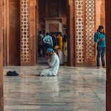 A man in white traditional attire and a blue cap is kneeling and praying on a polished marble floor, surrounded by intricately designed red sandstone walls. In the background, several people are standing, with one person leaning against a pillar and looking at a phone.