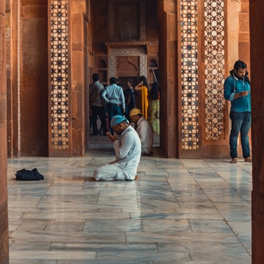 A man in white traditional attire and a blue cap is kneeling and praying on a polished marble floor, surrounded by intricately designed red sandstone walls. In the background, several people are standing, with one person leaning against a pillar and looking at a phone.
