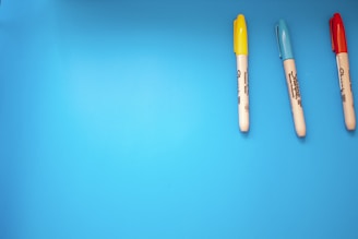 Three markers with yellow, blue, and red caps are aligned horizontally on a bright blue background.