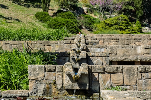 A well-designed stone structure built into a hillside features a series of zigzagging water channels. Green foliage, bushes, and a flowering tree add natural beauty to the setting above the stonework.