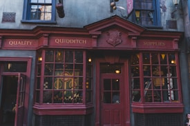 A storefront with vintage architecture featuring red wooden frames, large glass windows, and a sign reading 'Quality Quidditch Supplies'. The shop exhibits a medieval style with a coat of arms displayed above the door. Mannequins and various items appear in the window displays.
