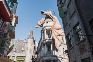 In a fantastical street setting, a large, winged dragon perched atop a building dominates the scene. The architecture has a medieval and whimsical style, featuring pointed roofs and stonework. The dragon appears menacing, with its mouth open, as if ready to breathe fire.