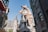 In a fantastical street setting, a large, winged dragon perched atop a building dominates the scene. The architecture has a medieval and whimsical style, featuring pointed roofs and stonework. The dragon appears menacing, with its mouth open, as if ready to breathe fire.