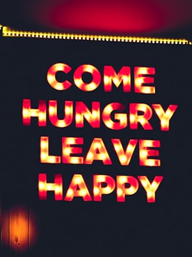 Brightly lit letters spell out the phrase 'COME HUNGRY LEAVE HAPPY' against a dark background. The letters have a vintage marquee style with warm, glowing lights around them.