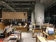 A modern bookstore interior with wooden shelves filled with an array of books and magazines. There are tables displaying various books with colorful covers. The space is well-lit, featuring overhead lights and a large window reflecting light from outside. Posters and signs with text are visible on the wall and some of the books are stacked vertically and horizontally.