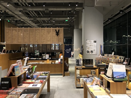 A modern bookstore interior with wooden shelves filled with an array of books and magazines. There are tables displaying various books with colorful covers. The space is well-lit, featuring overhead lights and a large window reflecting light from outside. Posters and signs with text are visible on the wall and some of the books are stacked vertically and horizontally.