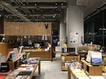 A modern bookstore interior with wooden shelves filled with an array of books and magazines. There are tables displaying various books with colorful covers. The space is well-lit, featuring overhead lights and a large window reflecting light from outside. Posters and signs with text are visible on the wall and some of the books are stacked vertically and horizontally.