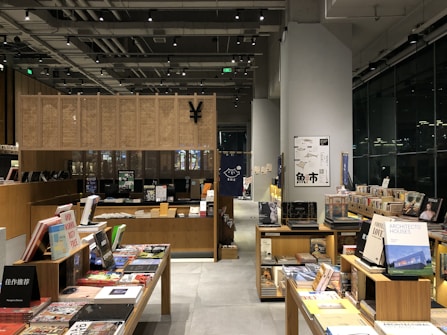 A modern bookstore interior with wooden shelves filled with an array of books and magazines. There are tables displaying various books with colorful covers. The space is well-lit, featuring overhead lights and a large window reflecting light from outside. Posters and signs with text are visible on the wall and some of the books are stacked vertically and horizontally.