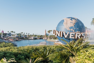 A large globe with the word 'Universal' prominently displayed in front. Lush greenery and palm trees surround the area, with a body of water and various buildings in the background, suggesting an amusement park setting. The sky is clear and bright, contributing to a lively atmosphere.