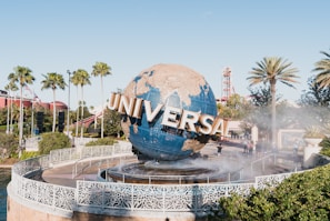 A large, metallic globe with the word 'UNIVERSAL' prominently displayed rotates on a fountain surrounded by a decorative railing. The background features palm trees, flags, and a backdrop of theme park elements such as roller coaster tracks. Clear blue skies indicate a sunny day.
