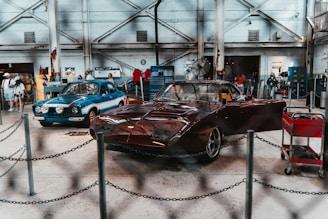 An indoor automotive exhibition featuring two classic cars. The car on the left is blue with a white stripe and additional lights on the front, while the car on the right is a red, vintage race car. Several people are present in the background, examining the cars and the surroundings. The space has an industrial feel with exposed beams, metal machinery, toolboxes, and an overall workshop ambiance.