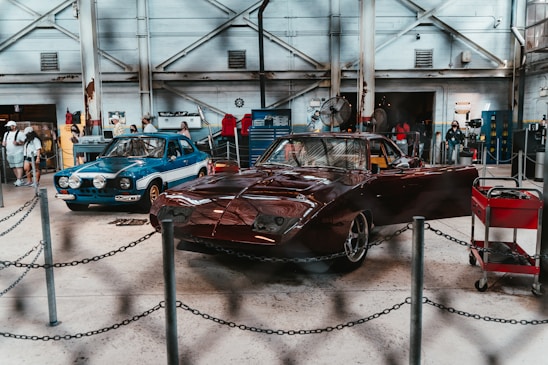 An indoor automotive exhibition featuring two classic cars. The car on the left is blue with a white stripe and additional lights on the front, while the car on the right is a red, vintage race car. Several people are present in the background, examining the cars and the surroundings. The space has an industrial feel with exposed beams, metal machinery, toolboxes, and an overall workshop ambiance.