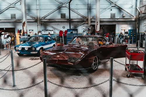 An indoor automotive exhibition featuring two classic cars. The car on the left is blue with a white stripe and additional lights on the front, while the car on the right is a red, vintage race car. Several people are present in the background, examining the cars and the surroundings. The space has an industrial feel with exposed beams, metal machinery, toolboxes, and an overall workshop ambiance.