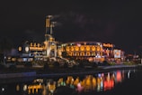 A nighttime scene featuring a brightly lit entertainment complex with two prominent buildings. The left structure has industrial-style elements with steam rising from chimneys. The right building, adorned with arches and illuminated signage, displays a 'Hard Rock' sign. The surrounding area includes manicured landscaping and a reflective body of water in the foreground, capturing the vibrant lights from the buildings.