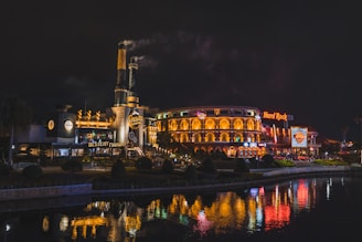A nighttime scene featuring a brightly lit entertainment complex with two prominent buildings. The left structure has industrial-style elements with steam rising from chimneys. The right building, adorned with arches and illuminated signage, displays a 'Hard Rock' sign. The surrounding area includes manicured landscaping and a reflective body of water in the foreground, capturing the vibrant lights from the buildings.