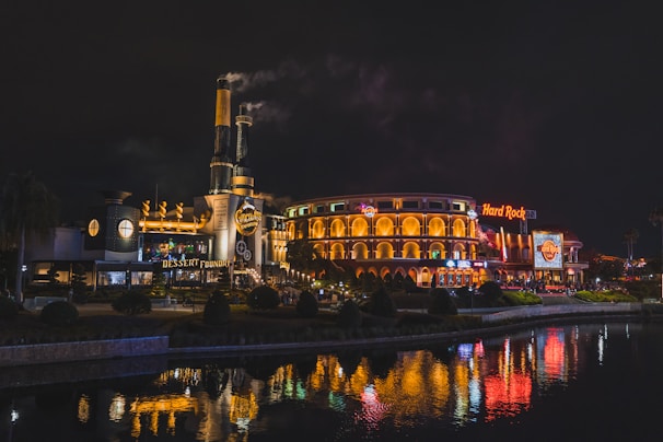 A nighttime scene featuring a brightly lit entertainment complex with two prominent buildings. The left structure has industrial-style elements with steam rising from chimneys. The right building, adorned with arches and illuminated signage, displays a 'Hard Rock' sign. The surrounding area includes manicured landscaping and a reflective body of water in the foreground, capturing the vibrant lights from the buildings.