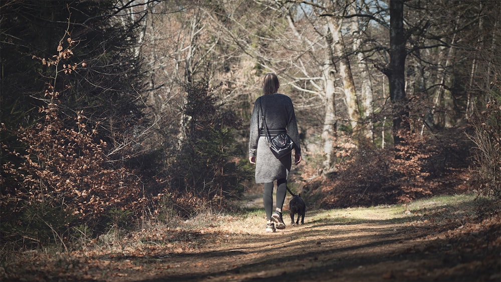 灰色のコートを着た女性が昼間、茶色の未舗装の道路を黒い犬と一緒に歩いている