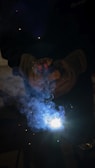 A welder is operating with protective gloves, and bright sparks are emitted from the welding process. Blue smoke surrounds the area, creating a dramatic effect with the intense white light at the center.