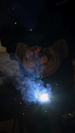 A welder is operating with protective gloves, and bright sparks are emitted from the welding process. Blue smoke surrounds the area, creating a dramatic effect with the intense white light at the center.